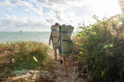 Two paddlers carrying inflatable SUPs in backpacks toward the ocean on a beach trail